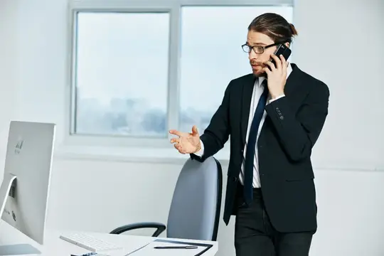 Мужчина в очках и костюме говорит по телефону у окна офиса