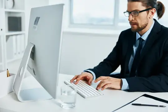 Мужчина в очках и деловом костюме работает за компьютером в светлом офисе