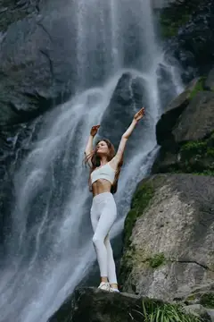 Молодая женщина с поднятыми руками стоит на камне у водопада