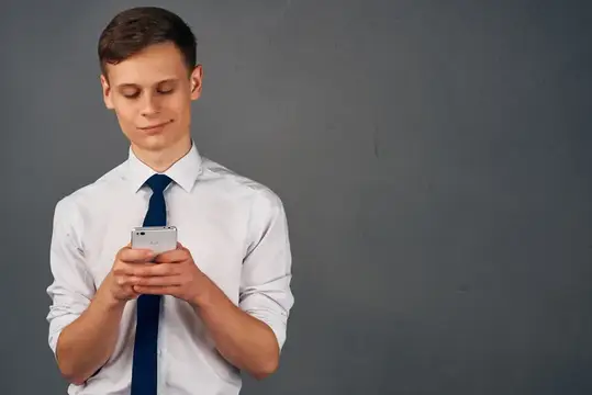 Молодой человек в белой рубашке смотрит в смартфон на сером фоне