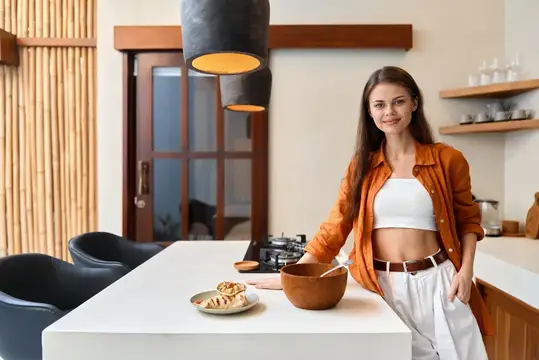 Улыбающаяся темноволосая женщина готовит на светлой кухне с деревянными полками
