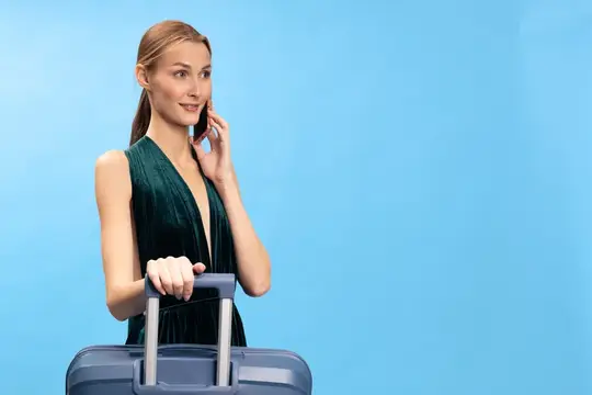 Улыбающаяся молодая женщина в зелёном платье держится за ручку чемодана и разговаривает по телефону на голубом фоне