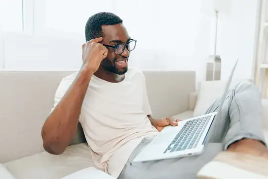 Улыбающийся темнокожий мужчина в очках работает за ноутбуком на диване