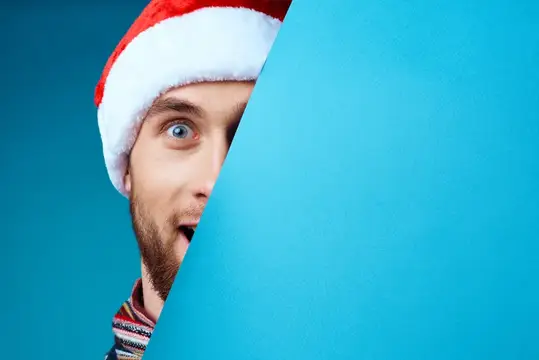 Удивлённый мужчина в новогодней шапке выглядывает из-за синей стены