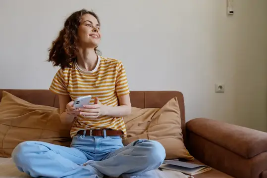 Улыбающаяся кудрявая девушка сидит на диване со смартфоном