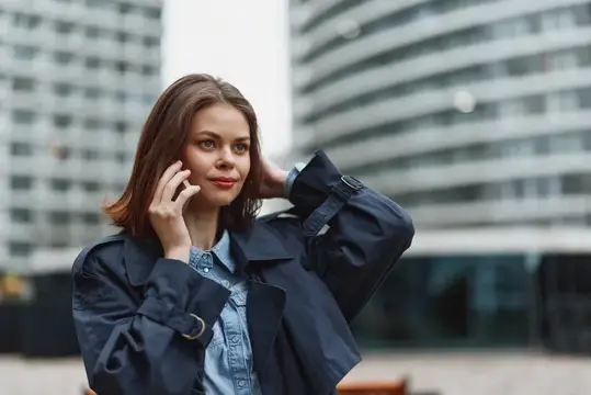 Крупный план, альтернативный ракурс той же сцены, подчёркивающий сосредоточенное выражение лица женщины во время разговора по телефону, видна джинсовая куртка, городской фон и современная атмосфера