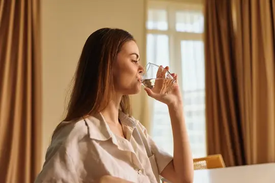 Молодая женщина пьёт воду из стакана у окна