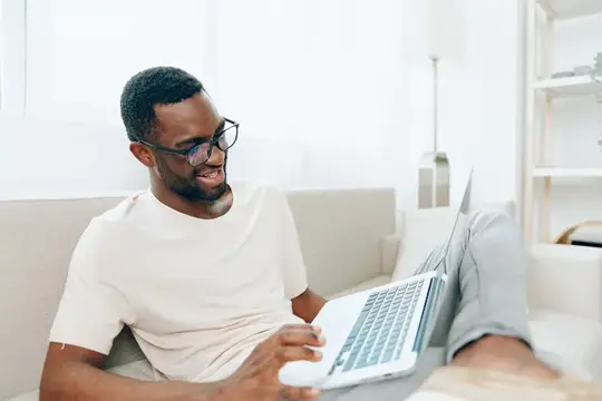Улыбающийся темнокожий мужчина в очках работает за ноутбуком на диване