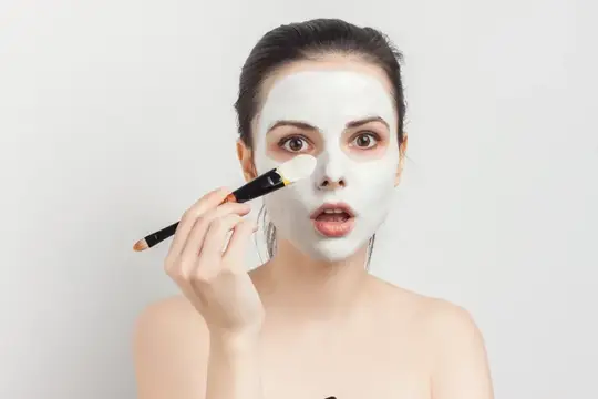 Молодая женщина наносит белую глиняную маску кисточкой на лицо