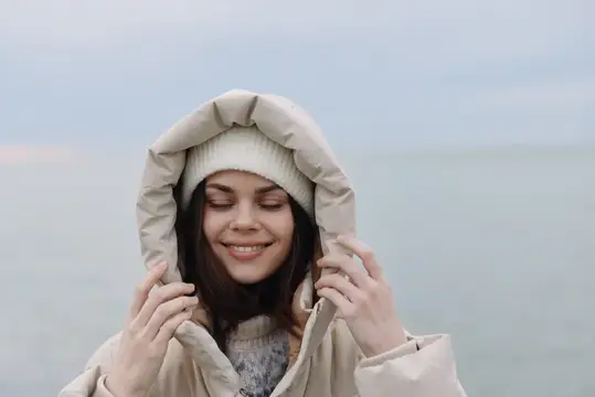 Улыбающаяся девушка в капюшоне и шапке на берегу моря