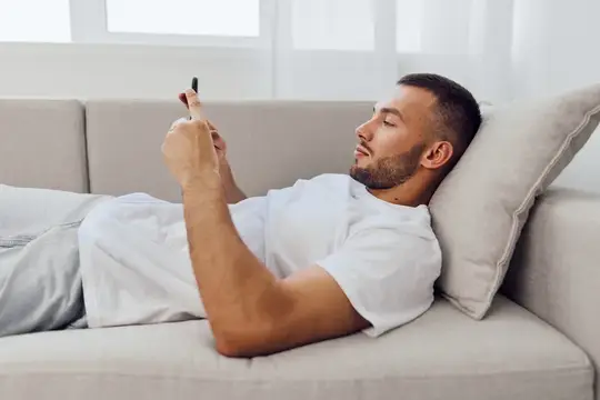 Молодой мужчина лежит на диване и смотрит в смартфон