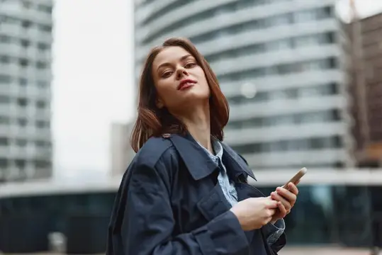 Молодая женщина в плаще с телефоном на фоне высоток