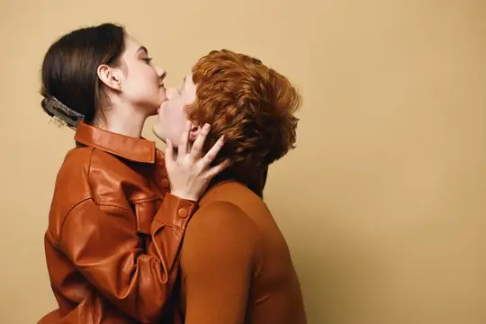 Две женщины в коричневых куртках нежно целуются на бежевом фоне