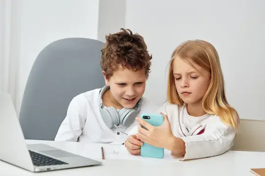 Мальчик и девочка смотрят в смартфон за столом с ноутбуком