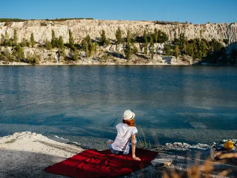 Рыжеволосая девушка в белой шляпе сидит на красном пледе у синего озера