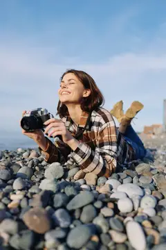 Портрет жизнерадостного фотографа на каменистом пляже: камера наготове, расслабленная поза, солнечный свет и морской бриз запечатлевают искренние прибрежные моменты