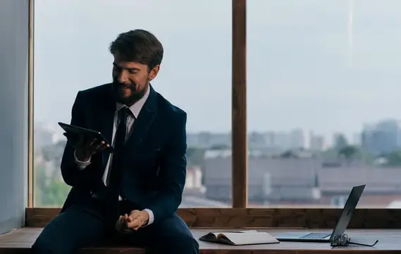 Улыбающийся бородатый мужчина в костюме с планшетом у панорамного окна