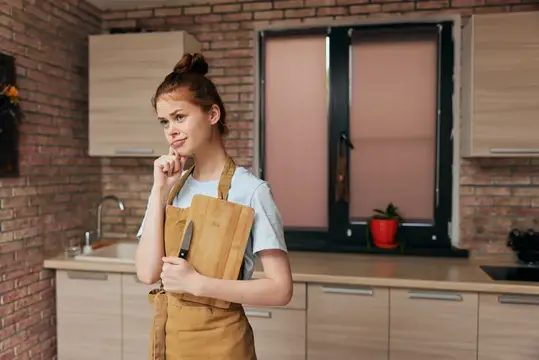 Задумчивая молодая женщина в фартуке с ножом и доской на кухне