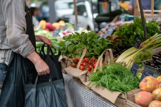 Покупатель с сумками выбирает свежие овощи и зелень на рынке
