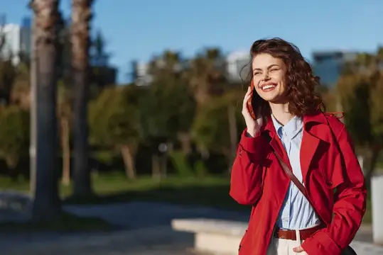 Улыбающаяся кудрявая девушка в красном плаще говорит по телефону на улице