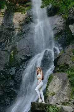 Девушка в белом стоит на камне у подножия водопада