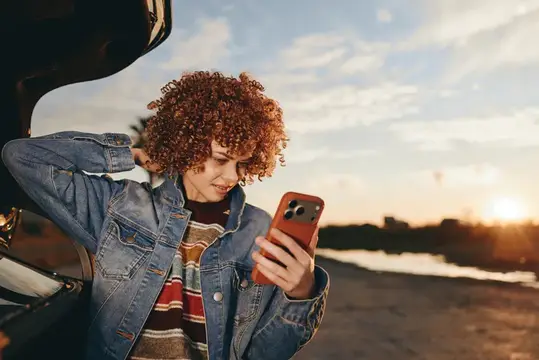 Женщина с вьющимися волосами делает селфи на смартфон в джинсовой куртке, улыбается на улице на закате