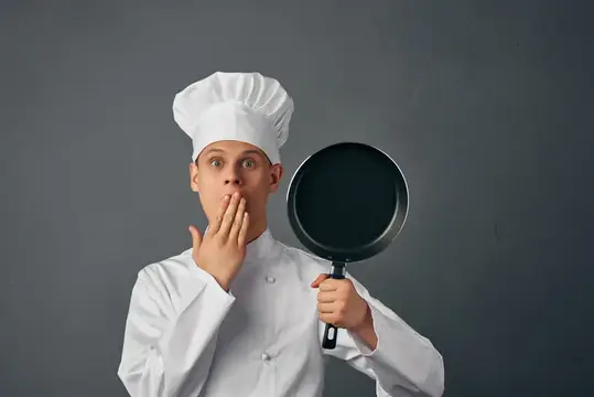 Удивлённый повар в колпаке с сковородой на сером фоне