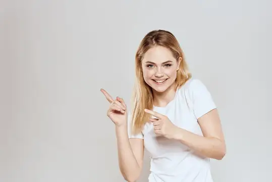 Улыбающаяся блондинка в белой футболке указывает пальцами в сторону