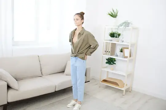 Молодая женщина в рубашке и джинсах стоит в светлой гостиной