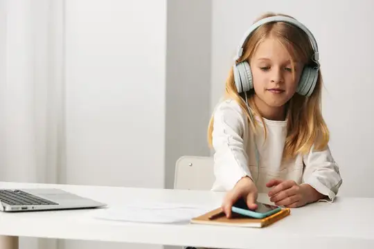 Светловолосая девочка в наушниках за столом с ноутбуком и смартфоном