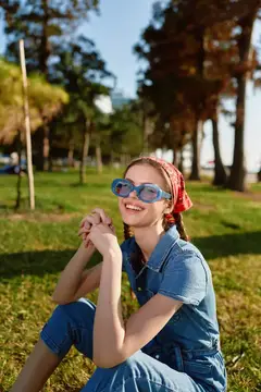 Улыбающаяся девушка в джинсовом комбинезоне и солнцезащитных очках сидит на траве в парке