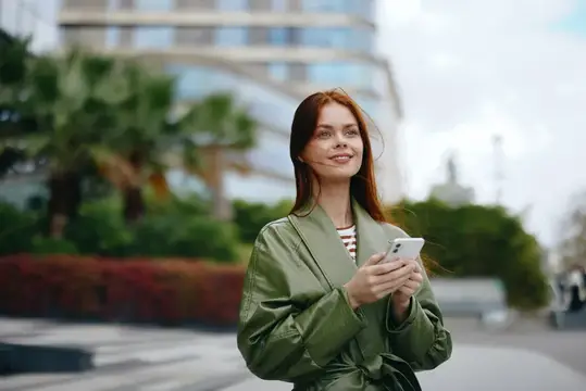 Деловая женщина с телефоном в руке, одетая в модную одежду, с улыбкой прогуливается по городу рядом с офисными зданиями