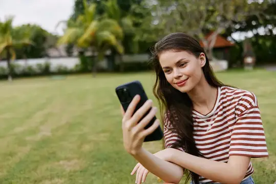 Улыбающаяся темноволосая девушка делает селфи на смартфон в парке