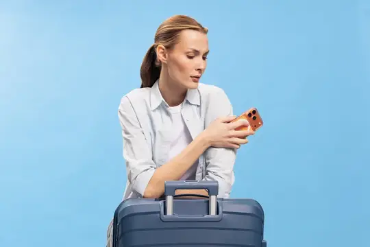 Сосредоточенная женщина со смартфоном в руке сидит за большим чемоданом на голубом фоне