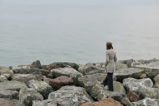 Мужчина в плаще и кепке стоит на камнях у моря