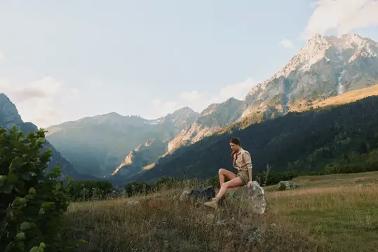 Молодая женщина, ведущая активный образ жизни, отдыхает на камне в горной долине, окруженная зеленой травой и вершинами, в солнечную погоду, в повседневной одежде, в спокойной и умиротворенной…
