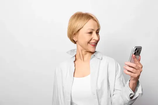 Улыбающаяся блондинка в белой рубашке смотрит в смартфон на светлом фоне