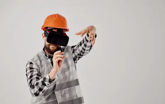 Бородатый мужчина в каске и VR-очках жестикулирует на сером фоне