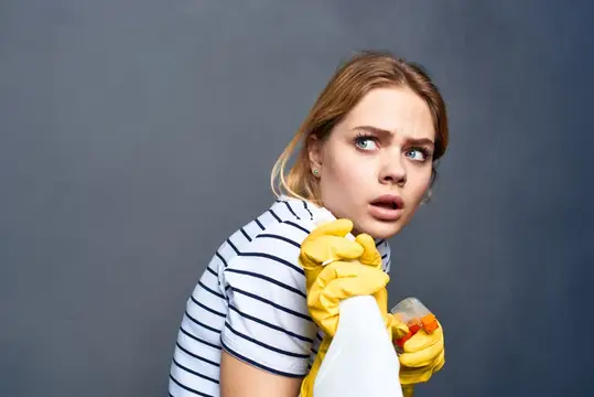 Удивлённая блондинка в жёлтых перчатках с бутылкой на сером фоне