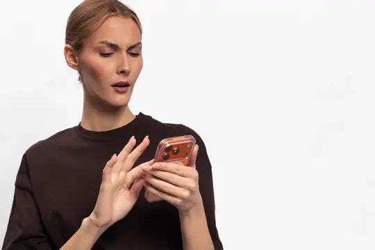 Удивлённая женщина в чёрной футболке смотрит в смартфон на белом фоне
