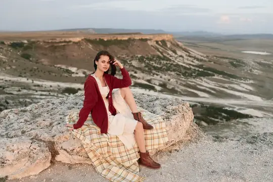 Молодая брюнетка в бордовом кардигане сидит на скалах в степи