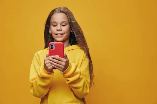 Улыбающаяся девочка с длинными волосами смотрит в красный смартфон на жёлтом фоне