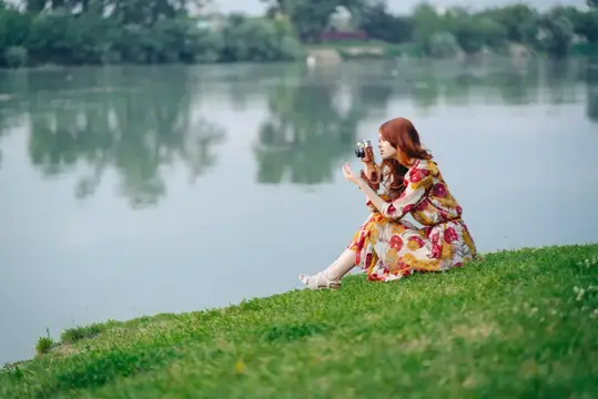 Рыжеволосая девушка в цветастом платье фотографирует у озера