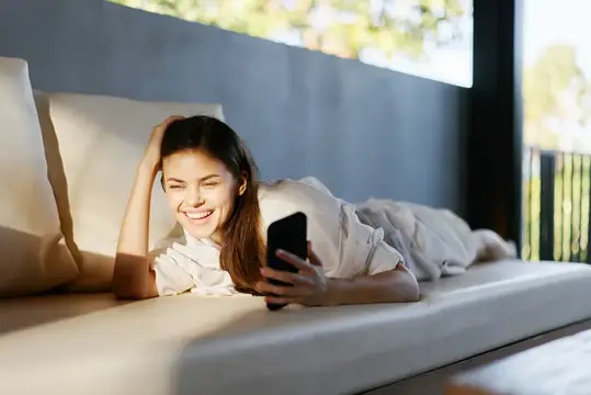 Улыбающаяся девушка лежит на кровати со смартфоном