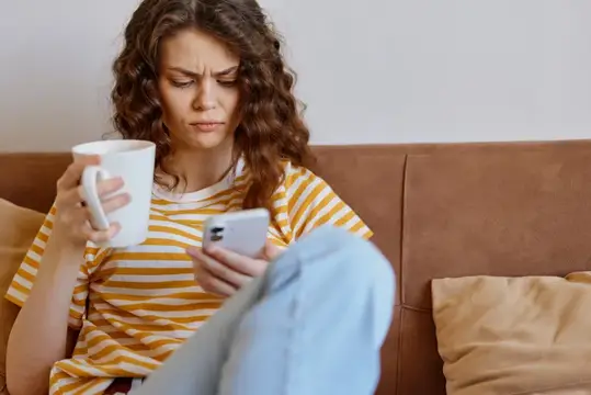 Кудрявая девушка с кружкой хмурится, глядя в телефон на диване