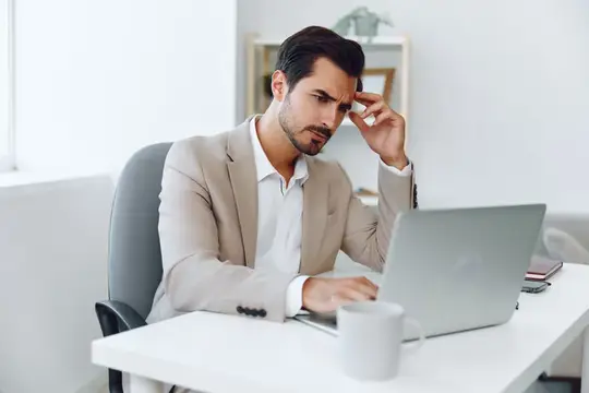 Уставший бородатый мужчина в светлом пиджаке работает за ноутбуком в офисе