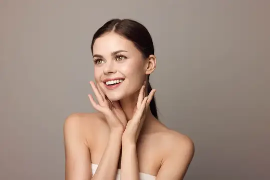 Улыбающаяся темноволосая женщина с ладонями у лица на сером фоне
