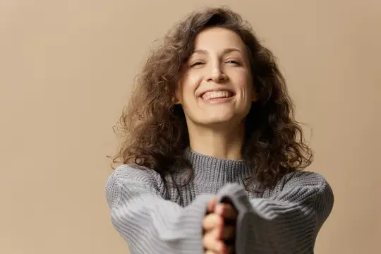 Дружелюбная, жизнерадостная, кудрявая красивая девушка в сером теплом повседневном свитере протягивает руки к камере, позирует изолированно на бежевом пастельном фоне