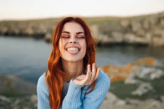 Рыжеволосая девушка с закрытыми глазами и высунутым языком на фоне моря