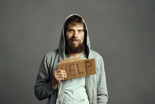 Бородатый мужчина в капюшоне держит картонный знак с надписью help